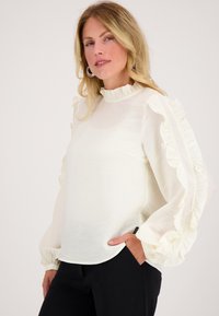 Witte blouse met rimpelige schouderdetails en lange, luchtige mouwen. Heeft een hoge halslijn en een zachte, gestructureerde stof. Gecombineerd met zwarte broeken.