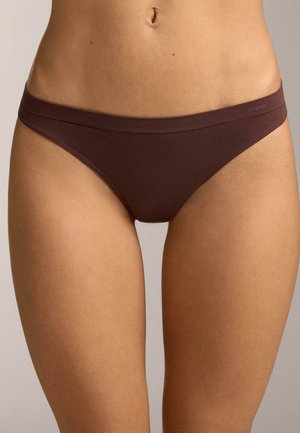 Női felsőtest, egy sima barna bikini stílusú fehérneműt visel, puha derékpánttal és minimális varrással, semleges háttér előtt.