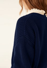 Osoba z jasnobrązowymi włosami ma na sobie granatowy dzianinowy sweter nałożony na białą bluzkę z falbankami, odwrócona plecami w kierunku gładkiego tła.