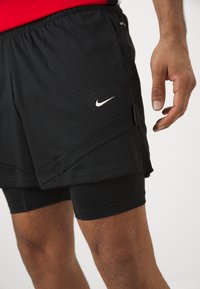 Pantalones cortos deportivos negros con textura de malla, con un logo blanco de Nike. Capa interior de pantalones cortos ceñidos negros visible por debajo.