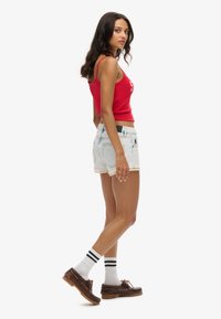 Top corto rojo, pantalones cortos de mezclilla claros con ribete de encaje, calcetines blancos con rayas negras y zapatos marrones. El modelo está de perfil.