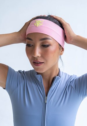 Pink fabric headband with a yellow geometric logo, set against a light grey background. Model wears a fitted light blue zip-up athletic top.