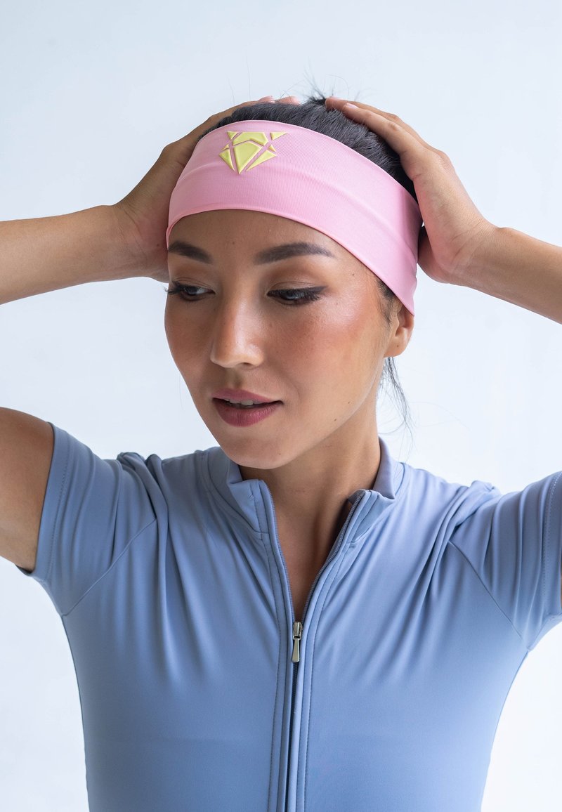 Pink fabric headband with a yellow geometric logo, set against a light grey background. Model wears a fitted light blue zip-up athletic top.