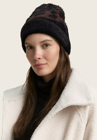 Schwarze und braune gemusterte Strickmütze mit einer flauschigen Textur, versehen mit einem gerippten Saum. Getragen mit einem weißen Wollmantel und einem schwarzen Rollkragenpullover.