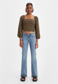 Kobieta stoi w czarnej, kwiecistej crop topie z kwadratowym dekoltem i bufiastymi rękawami, w jasnoniebieskich dzwonach i czarnych sandałach na białym tle.