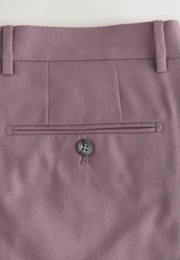 Tylna kieszeń fioletowo-różowych spodni wizytowych z pojedynczym czarnym guzikiem i dwoma pętelkami na pasek widocznymi na talii.