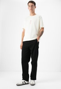 A young man stands confidently, wearing a light-colored t-shirt with a floral design, black pants, and casual sneakers on a white background.