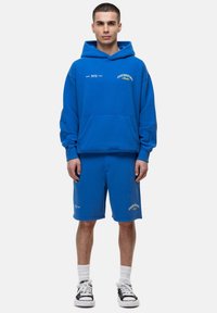 Felpa blu con tasca frontale, abbinata a pantaloni corti coordinati. Presenta accenti di testo bianchi e dorati. Indossata con calze bianche e scarpe nere.
