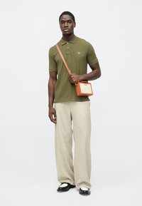 Πόλο μπλούζα ελιάς, κρεμ παντελόνι με φαρδιά μπατζάκια, μαύρα και λευκά παπούτσια, και πορτοκαλί τσάντα χιαστί με δομημένο σχέδιο.