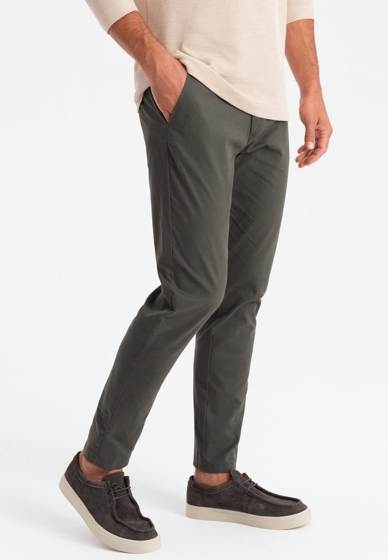 Homme portant un pantalon slim vert foncé et des baskets décontractées marron foncé, une main dans la poche, debout devant un fond uni.