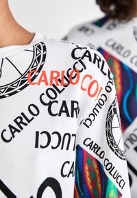 Bílá látka s opakujícími se nápisy "CARLO COLUCCI" v černé a oranžové barvě a duhovými vzorovanými detaily na oblečení dvou osob.