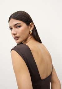 Brown dress with a deep square back, slim straps, and smooth texture. Gold hoop earrings add contrast and shine. Neutral background.