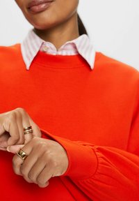 Sudadera naranja con cuello de canalé y mangas abullonadas, superpuesta a una camisa de rayas en rosa claro y blanco. Anillos dorados en las manos.