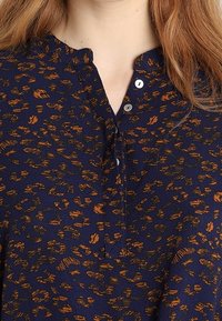 Granatowa bluzka z abstrakcyjnym pomarańczowym wzorem, z luźnym kołnierzem i dwoma metalowymi guzikami przy dekolcie. Miękka faktura materiału.