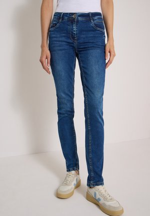 Person iført slim fit blå jeans og hvide sneakers med beige såler, stående mod en ensfarvet lys baggrund.
