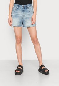 Light blue denim shorts with a distressed design, frayed hem, and five-pocket style, paired with black buckle sandals on a neutral background.