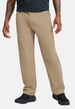 Uomo con pantaloni beige a gamba dritta e scarpe da ginnastica nere, in piedi davanti a uno sfondo bianco semplice.