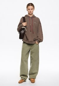 Sudadera marrón de gran tamaño con cordones rojos y blancos, combinada con pantalones holgados verde oliva y zapatos de ante marrones. Lleva un bolso de cuero oscuro en la mano.