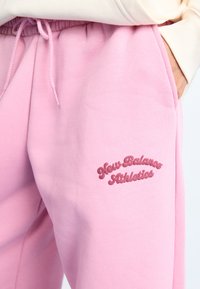 Pantalon de survêtement rose en tissu doux, présentant une taille élastique et un logo "New Balance Athletics" cousu en rose plus foncé.