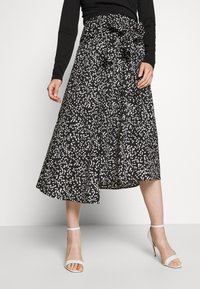 Kvinna som bär en svart och vit prickig midi-kjol med en knuten midjebow, svart långärmad topp och vita högklackade sandaler.