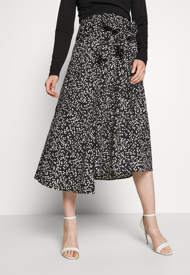 Kvinna som bär en svart och vit prickig midi-kjol med en knuten midjebow, svart långärmad topp och vita högklackade sandaler.