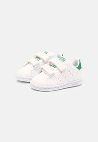 White athletic shoes with green accents, featuring dual Velcro straps, perforated details on the sides, and smooth synthetic material.