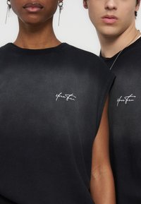 Deux personnes portant des hauts noirs sans manches avec un texte blanc en cursive sur la poitrine, montrant des boucles d'oreilles et un collier.