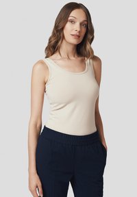 Beżowy top na ramiączkach o prążkowanej teksturze z okrągłym dekoltem, zestawiony z ciemnonawowymi spodniami na elastycznej talii, na szarym tle.