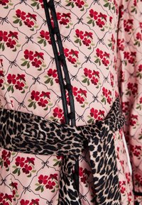 Το φλοράλ φόρεμα διαθέτει ροζ φόντο με κόκκινα λουλούδια και πράσινα φύλλα, διακοσμημένο με μαύρα κουμπιά και ζωνάκι με λεοπάρ σχέδιο.