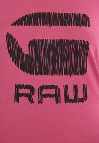 Pink bomulds T-shirt med et stort sort doodle-stil grafik, der viser teksten "RAU" med teksturerede linjer og ujævne kanter.