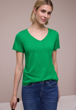 Młoda kobieta w zielonej koszulce z dekoltem w serek i niebieskich dżinsach, stojąca na jednolitym beżowym tle, delikatnie się uśmiechając.