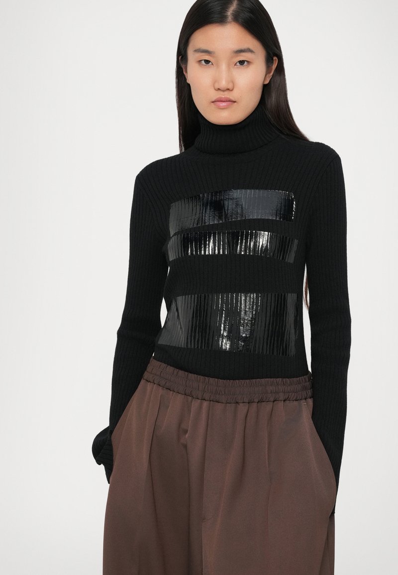 Pull à col roulé côtelé noir avec trois bandes horizontales brillantes noires. Associé à un pantalon élastique marron. Fond uni.