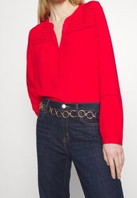 Blouse rouge à manches longues avec un col en V et un devant texturé, associée à un jean bleu foncé et une ceinture en chaîne circulaire dorée.