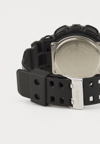 Montre G-Shock noire avec un bracelet en caoutchouc texturé, un fond en acier inoxydable et une boucle en métal argenté. Ses caractéristiques incluent plusieurs trous dans le bracelet.