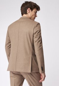 Hellbrauner taillierter Blazer aus strukturiertem Stoff, mit einem Schlitz, Latzschlägen und schwarzen Knopfakzenten an den Ärmeln.