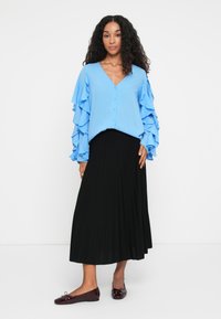 Blouse bleu clair avec des manches volantées, jupe noire plissée et ballerines bordeaux. La blouse a un col en V et une fermeture à boutons.