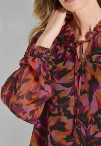 Durchsichtige, mehrfarbige Bluse mit einem lebhaften Blumenmuster in Rosa-, Orange- und Schwarztönen. Rüschenausschnitt und gepuffte Ärmel mit Knopfakzenten.