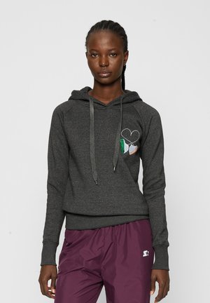 Žena s vrkočmi, ktorá nosí tmavosivú mikinu s kapucňou s motívom srdca a tenisky, v kombinácii s bordovými športovými nohavicami na jednofarebnom pozadí.