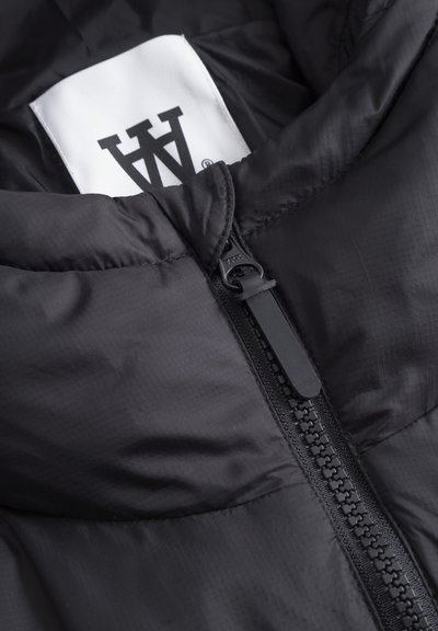 Doudoune noire avec un col haut, dotée d'une fermeture éclair bien visible et d'un tissu lisse et brillant. L'étiquette intérieure affiche le logo de la marque.