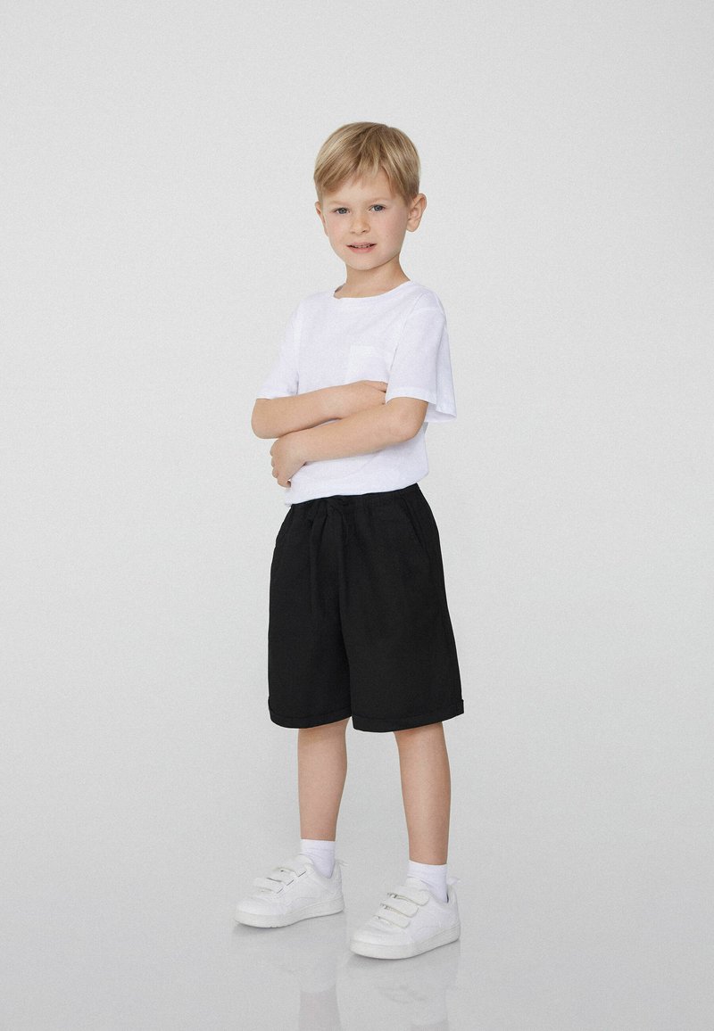 Bílé tričko s krátkými rukávy a kapsou, zkombinované s černými šortkami do délky kolen. Bílé tenisky a viditelné kotníkové ponožky.