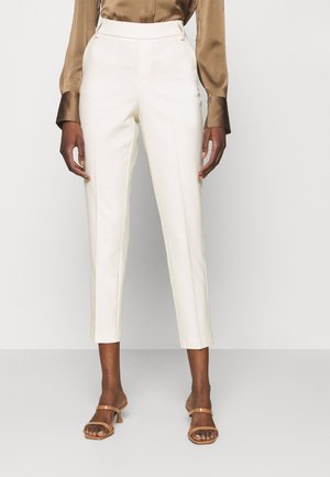 Frau trägt cremefarbene, maßgeschneiderte Ankle-Hosen, braune Satinbluse und beige Sandaletten mit Absatz, steht vor einem einfarbigen Hintergrund.