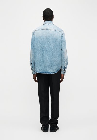 Άνδρας όρθιος με την πλάτη στραμμένη, φορώντας ανοιχτό μπλε τζιν μπουφάν, μαύρο παντελόνι και μαύρα αθλητικά παπούτσια Versace, μπροστά σε απλό λευκό φόντο.