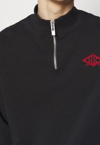 Black pullover with a high collar, featuring a silver zip and red embroidered logo. Soft texture and minimalistic design.