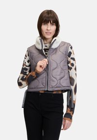 Gepolsterte graue Weste mit einem Kunstfellkragen, Frontreißverschluss und kürzem Design. Das Model trägt darunter einen gemusterten Pullover. Schwarze Hosen vervollständigen das Outfit.