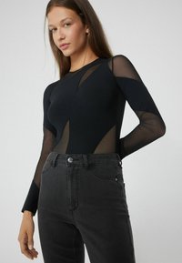 Mladá žena oblečená v černém topu s dlouhým rukávem a průsvitnými síťovanými panely, v trendech vysokého pasu, tmavě šedé džíny, pózující na jednolitém pozadí.