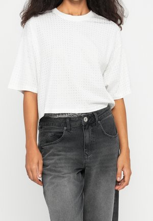 Vrouw draagt een wit gebreid topje met korte mouwen en kleine gaatjes, gecombineerd met een hooggetailleerde, vervaagde zwarte jeans die een geruite tailleband toont.