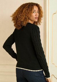 Crna majica s rebrastim tijelom, teksturiranim kabelskim pletenim rukavima i resastim rubom. Gladak materijal, duži rukavi i prilagođen silueta.