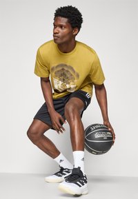 Gul grafisk t-skjorte, svarte shorts, hvite sokker, sort og hvit sneakers. Holder en basketball, i en knebøyposisjon, nøytral bakgrunn.