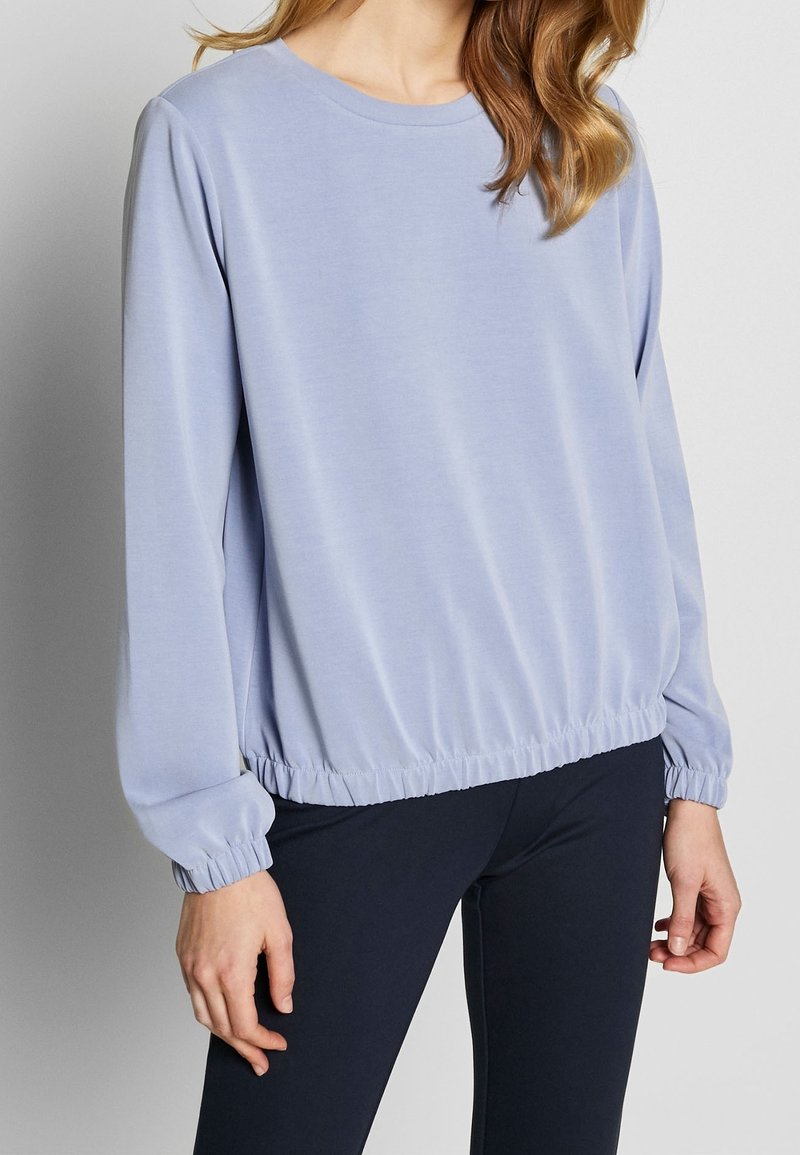 Femme portant un haut à manches longues bleu clair avec des poignets et un ourlet élastiques, associé à un pantalon foncé ajusté, sur un fond uni.