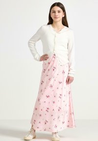 Witte lange mouwen cardigan met knopen over een roze bloemen maxi-jurk met kleine rozen en groene bladeren, gecombineerd met witte sneakers.